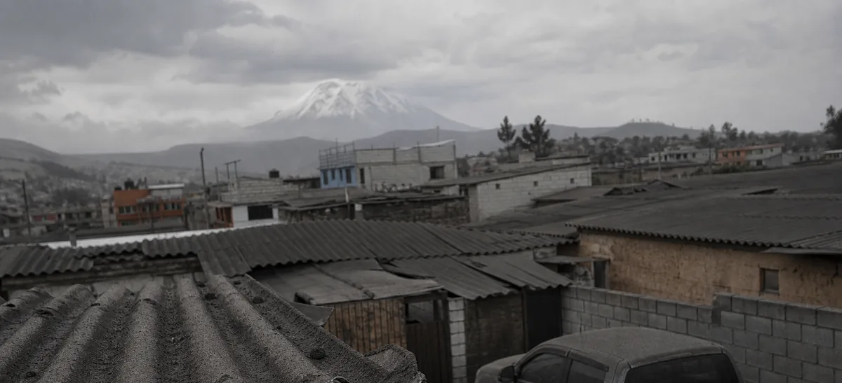 Ceniza volcánica gris depositada sobre los techos de zinc y una camioneta de un pueblo andino, con un volcán nevado difuso al fondo.
