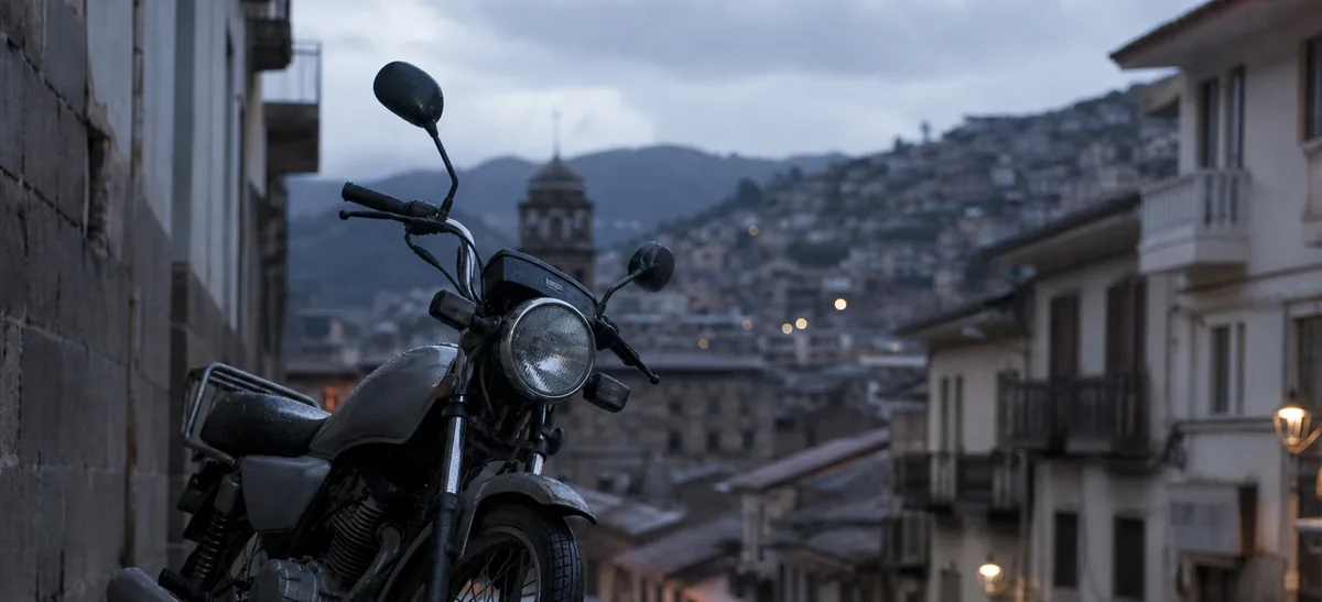 Una motocicleta de baja cilindrada aparcada en una calle empedrada mojada al atardecer en una ciudad andina.