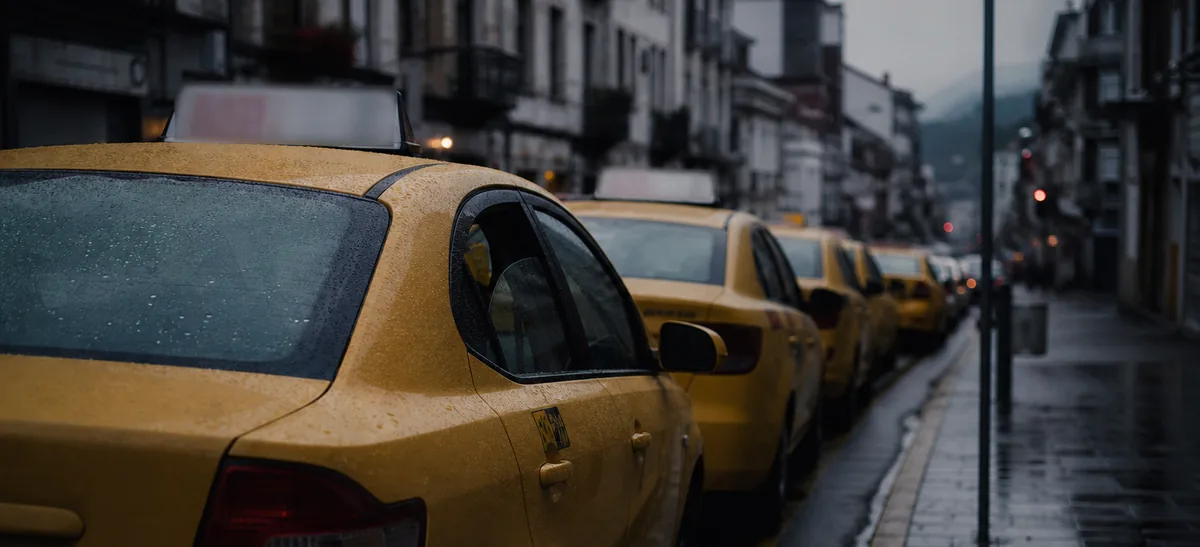Una fila de taxis amarillos esperando en una parada de una calle mojada en Ecuador, vista desde la acera con poca luz.