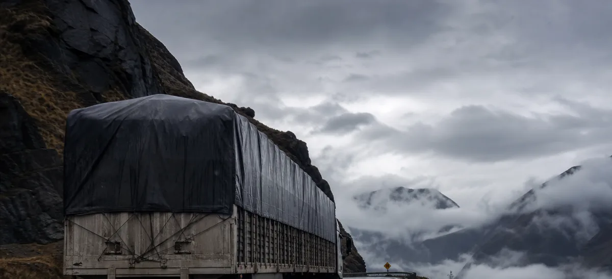 Un camión articulado cargado en una carretera de montaña mojada en los Andes, junto a un quitamiedos y un valle con nubes.